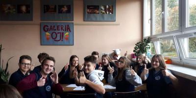 Grupa uczniów siedzi w klasie i uśmiechnięta pokazuje kciuki w górę.
